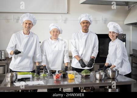 Portrait eines jungen multirassischen Teams von Köchen, die in der professionellen Küche Essen zum Mitnehmen zubereiten. Konzept der dunklen Küche zum Kochen für die Lieferung. Idee der Teamarbeit in einem Restaurant. Vielfältige Menschen am Arbeitsplatz Stockfoto