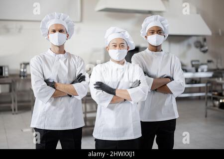 Porträt eines Teams aus drei verschiedenen Rassen, die in der professionellen Küche zusammenstehen. Gut gekleidete Köche in Gesichtsmasken und Schutzhandschuhen bereit für einen Job. Neue Normalität für das Geschäft während einer Pandemie Stockfoto