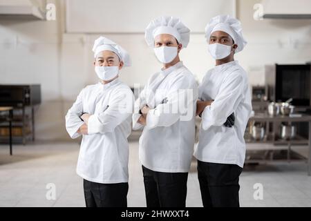 Porträt eines Teams aus drei verschiedenen Rassen, die in der professionellen Küche zusammenstehen. Gut gekleidete Köche in Gesichtsmasken und Schutzhandschuhen bereit für einen Job. Neue Normalität für das Geschäft während einer Pandemie Stockfoto