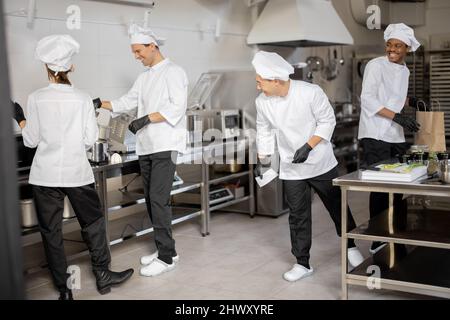 Fröhliches multirassisches Team von Köchen arbeitet aktiv in der Küche. Der asiatische Koch kündigt die Bestellliste vom gedruckten Scheck an, der lateinische Kerl verpackt Lebensmittel für die Lieferung, die europäischen Köche kochen hinterher. Konzept der Teamarbeit im Restaurant Stockfoto