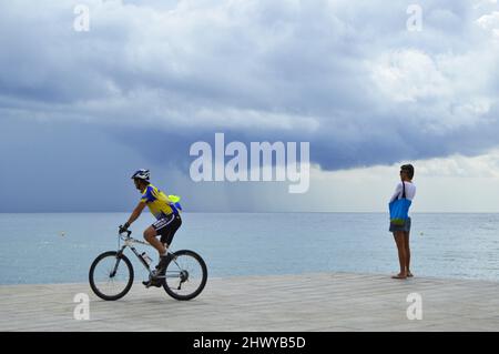 Menschen auf dem gepflasterten Weg am Strand von Barceloneta, stürmische Wolken nähern sich der Küste von Barcelona Spanien. Stockfoto