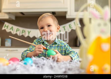 Netter kleiner Junge, der Ostereier in der Küche dekoriert Stockfoto