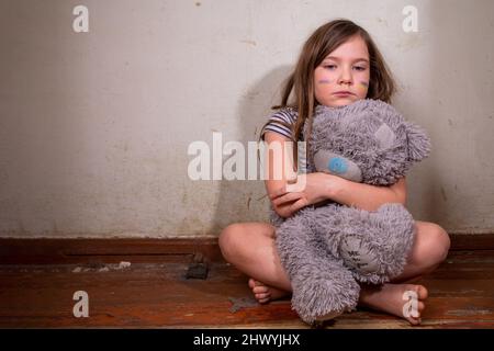 Stop Krieg in der Ukraine Konzept. Kleines ukrainisches blondes Mädchen, ukrainische Flaggen auf Wangen gemalt, mit Teddybären-Spielzeug in einem ruinierten alten Haus. Rette das Chill Stockfoto