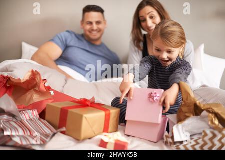 Oh wow, danke Mama und Papa. Aufnahme eines kleinen Mädchens, das Geschenke im Bett öffnet, während ihre Eltern zusehen. Stockfoto