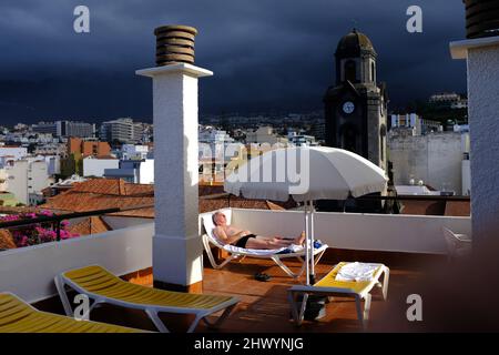 Ein älteres Männchen, das auf einem Sonnenbett mit der Stadt Puerto de la Cruz, Teneriffa, im Hintergrund, Stockfoto