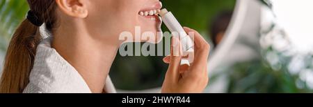 Seitenansicht einer lächelnden Frau im Bademantel, die Lippenbalsam aufbringt, sich um ihre Lippen kümmert und im Badezimmer steht Stockfoto