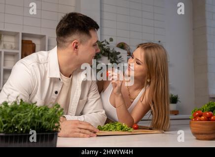 Glückliches Paar, das zu Hause kocht. Schöne Frau füttert Ehemann Tomate, während Gemüse Salat. Gesunde Ernährung Konzept. Zärtliche Beziehung zwischen verheirateten Menschen. Hochwertige Fotos Stockfoto