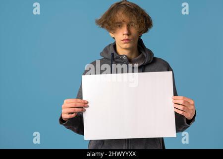 Ein ernsthafter, verärgerter Teenager mit grauem Hoodie und welligen Haaren hält ein leeres Poster auf blauem Hintergrund. Das Konzept des Protests. Modell Stockfoto