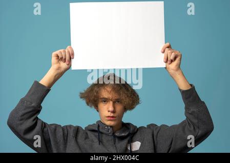 Ein ernsthafter, verärgerter Teenager mit grauem Hoodie und welligen Haaren hält ein leeres Poster auf blauem Hintergrund. Das Konzept des Protests. Modell Stockfoto