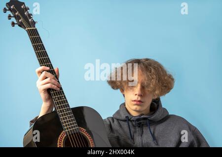 Ein Teenager in einem grauen Hoodie und welligen Haar hält eine Gitarre auf einem blauen Hintergrund. Platz für Text Stockfoto