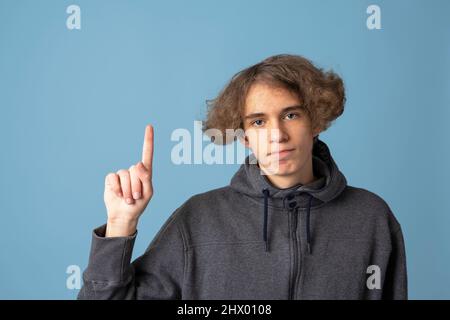 Ein Teenager in einem grauen Hoodie mit welligen Haaren zeigt mit seinem Zeigefinger auf einen blauen Hintergrund, Kopierfläche Stockfoto