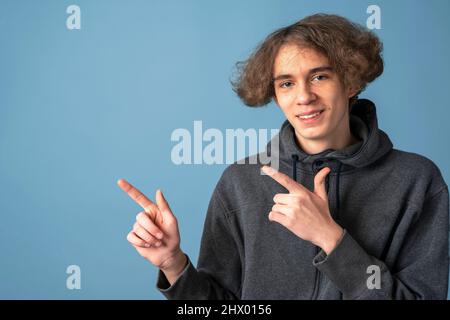 Ein lächelnder Teenager in einem grauen Hoodie und welligen Haaren zeigt mit den Händen auf einen blauen Hintergrund, schaut auf die Kamera Stockfoto