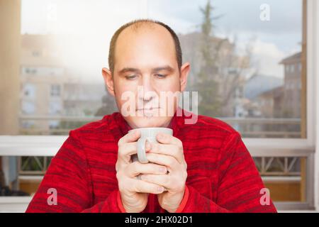 Ein kahler Mann in einem roten Pullover hält eine Tasse heißen, dampfenden Kaffee in beiden Händen bereit, um ihn zu trinken. Im Hintergrund ist ein Fenster und man kann Buildin sehen Stockfoto