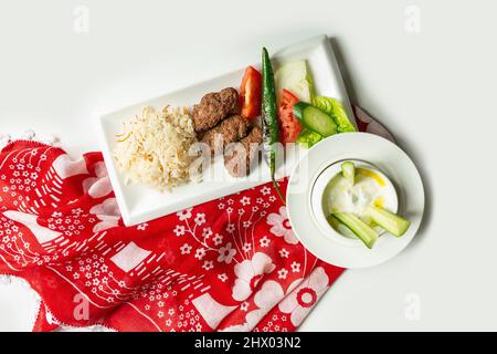 Traditionelle türkische Küche Izgara Kofte wuth Reis, Raita und Salat in einem Gericht isoliert auf bunten Tischtuch Draufsicht auf grauem Hintergrund Stockfoto
