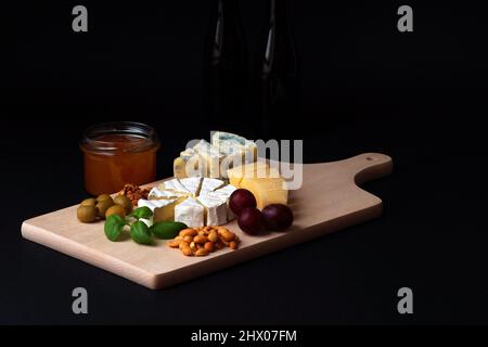 Auf schwarzem Hintergrund wurde eine Käseplatte, die auf ein Holzbrett gelegt wurde, zusammen mit Snacks - Oliven, Nüsse, Trauben und Basilikumblättern, n, fotografiert Stockfoto