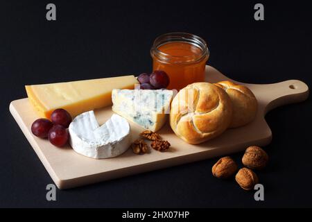 Auf dem Brett liegen drei Käsesorten - Camembert, Blau und Parmesan. Garniert mit Honig, Nüssen und Trauben. Es gibt auch Weizenbrötchen. Stockfoto