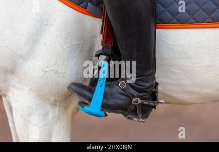 Der Fuß des Reiters, sitzend auf einem weißen Pferd, in einem schwarzen Stiefel mit einem Sporn, ruht auf einem sicheren Kunststoffbügel. Nahaufnahme. Reitturniere zeigen Stockfoto