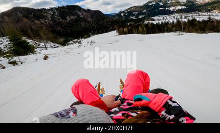 Eltern, die ein Kind auf einem Schlitten fahren, sich schnell auf der holprigen Schneestrecke bewegen, aus der Perspektive aufgenommen. Stockfoto