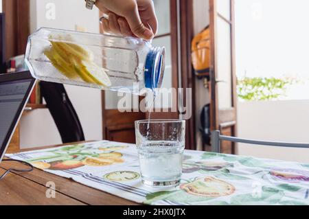 Nahaufnahme einer kaukasischen weiblichen Hand, die Limonade in einem Glas auf den Tisch gießt, mit der Eingangstür und natürlichem Licht von der Straße im Hintergrund. Stockfoto