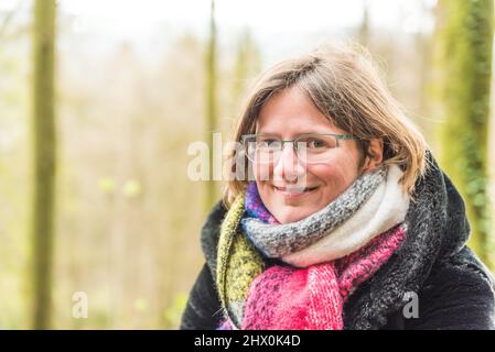 Porträt einer dreißigjährigen weißen Frau mit einem bunten Schal, einer Brille und einem grünen Bokeh-Naturhintergrund, Deutschland Stockfoto