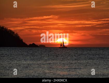 Ein spektakulärer orangefarbener Sonnenuntergang mit wirbelnden Wolken und der Silhouette eines Segelbootes vor der Sonne in Potrero Costa Rica. Stockfoto