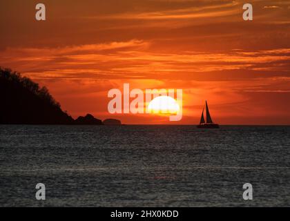 Ein spektakulärer orangefarbener Sonnenuntergang mit wirbelnden Wolken und der Silhouette eines Segelbootes, das in Potrero Costa Rica in Richtung Sonne segelt. Stockfoto