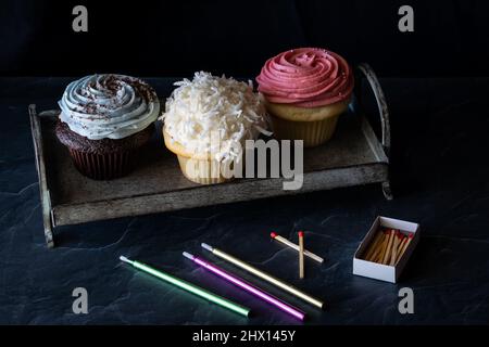 Ein Metalltablett mit drei dekorierten Cupcakes mit Kerzen und Streichhölzern davor. Stockfoto