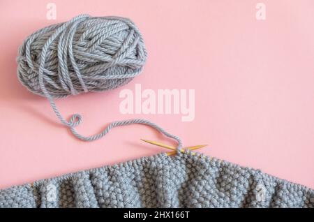 Stück graues Strickgewebe auf den Nadeln mit Garnknäuel, Strickverfahren auf rosa Hintergrund. Platz für Text kopieren .Draufsicht flache Lay-Vorlage Stockfoto