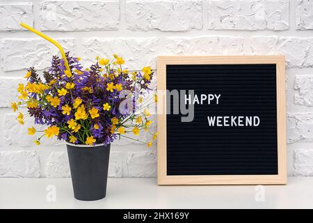 Happy Weekend Worte auf schwarzem Briefbrett und Strauß von farbigen Blumen in schwarzem Papier Kaffee Tasse mit Cocktail Stroh auf dem Tisch gegen weißen Ziegel wa Stockfoto