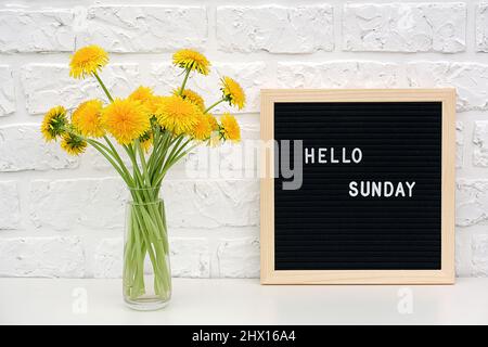 Hallo Sonntag Worte auf schwarzem Briefbrett und Bouquet von gelben Dandelions Blumen auf dem Tisch gegen weiße Ziegelwand. Konzept Happy Monday. Vorlage für Stockfoto