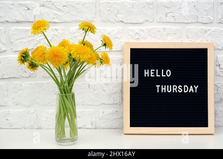 Hallo Donnerstag Worte auf schwarzem Briefbrett und Strauß von gelben Dandelions Blumen auf dem Tisch gegen weiße Ziegelwand. Konzept Happy Monday. Vorlage Stockfoto