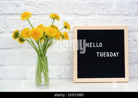 Hallo Dienstag Worte auf schwarzem Briefbrett und Strauß von gelben Dandelions Blumen auf dem Tisch gegen weiße Ziegelwand. Konzept Happy Monday. Vorlage f Stockfoto