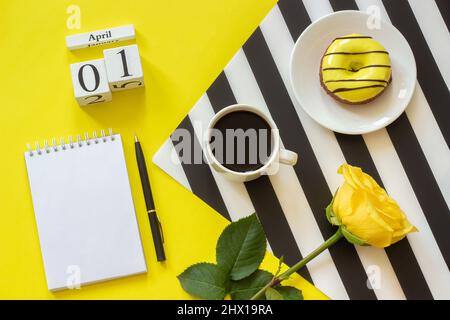 Holzwürfel Kalender April 1.. Tasse Kaffee, gelber Donut und Rose auf schwarz-weißer Serviette, leerer offener Notizblock für Text auf gelbem Hintergrund. Kontra Stockfoto