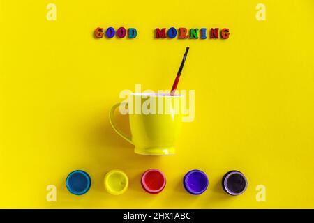 Schriftzug Good Morning aus bunten Buchstaben, Gläsern Gouache und gelber Kaffeetasse mit Pinsel. Stillleben auf gelbem Hintergrund Flat Lay Draufsicht, g Stockfoto