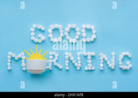 Text „Guten Morgen“ aus dem Marschlund und Kaffeetasse mit gelber aufgehender Sonne auf blauem Hintergrund. Konzept Stockfoto