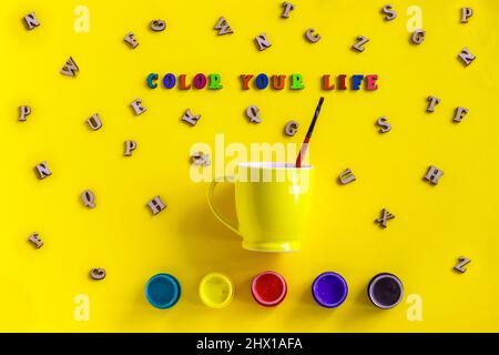 Text Farbe Ihr Leben aus bunten Buchstaben, Gouache, gelbe Kaffeetasse mit Pinsel, verstreute Holzbuchstaben auf gelbem Hintergrund Draufsicht, Greeti Stockfoto