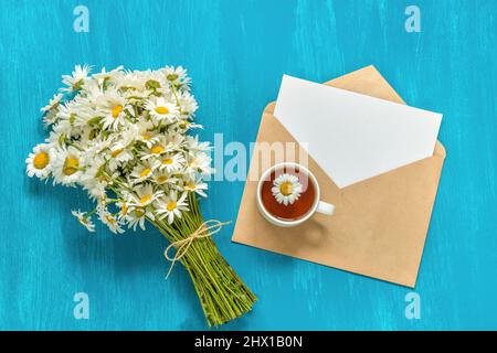 Blumenstrauß weiße Kamillen Blumen Tasse Kräuter Tee und Brief Handwerk Umschlag auf blauem Hintergrund rustikal vintage Konzept Good Morning Summer Tea Time GRE Stockfoto