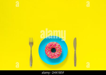 Pinker Donut auf blauem Teller und Messerschnittelgabel. Stillleben auf gelbem Hintergrund. Copy Space Draufsicht Flat Lay No Diet Day Stockfoto