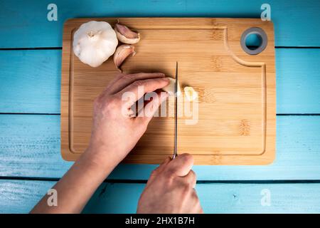 Die Hände der Frau schneiden mit einem Messer Knoblauch auf einem hölzernen Schneidebrett. Stockfoto