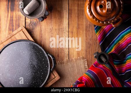 Mexikanische Küche Kochgeschirr. Bunte traditionelle Stoffe, comal de peltre, olla de barro und Molcajete auf rustikalem Holztisch. Draufsicht mit Kopierbereich Stockfoto