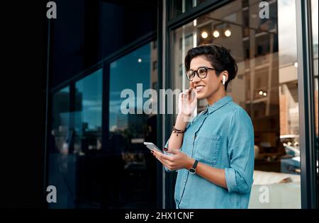 Musik hilft mir beim Warten. Ausgeschnittene Aufnahme einer attraktiven Frau, die draußen steht und ihre Ohrhörer einlegt, um über ihr Mobiltelefon Musik zu hören. Stockfoto