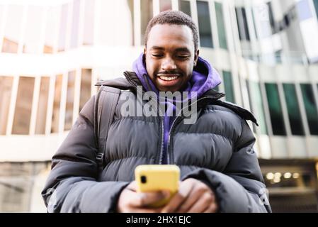 Von unten fröhlicher afroamerikanischer Mann in Oberbekleidung liest und sendet SMS auf dem Smartphone, während er auf der modernen Stadtstraße steht Stockfoto