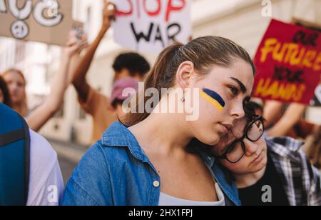 Beendet den Konflikt in der Ukraine. Junge Frau, die gegen den Krieg marschiert, mit der Flagge der Ukraine auf ihrem Gesicht. Gruppe von multikulturellen jungen Menschen holdin Stockfoto