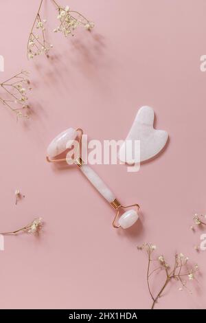 Rosenquarz-Massage-Rolle auf rosa Hintergrund mit Gypsophila-Blüten. Massagegeräte mit Jadestein, Anti-Aging, Anti-Falten-Beauty Skincar Stockfoto