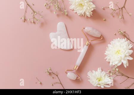 Rosenquarz-Massage-Rolle auf rosa Hintergrund mit Gypsophila-Blüten. Massagegeräte mit Jadestein, Anti-Aging, Anti-Falten-Beauty Skincar Stockfoto