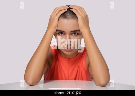 Das kaukasische Mädchen mit kurzen Haaren, fast kahl, hält ihre Hände hinter ihrem Kopf und lehnt sich in einem orangefarbenen T-Shirt auf einem hellen Hintergrund in Nahaufnahme alarmierend auf den Tisch. Das Konzept von Krankheit und Einsamkeit Stockfoto