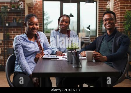 Happy african american Business Company Team sitzt am Schreibtisch in modernen Büroeinrichtung. Professionelle Kollegen der Marketingagentur sitzen am Tisch, lächeln und blicken auf die Kamera. Stockfoto