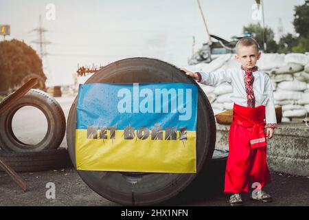Ukrainischer Junge in traditioneller Nationaltracht steht auf Straßensperre in der Nähe von Reifen mit Inschrift vor dem Hintergrund von Sandsäcken. Konzept der russischen mi Stockfoto