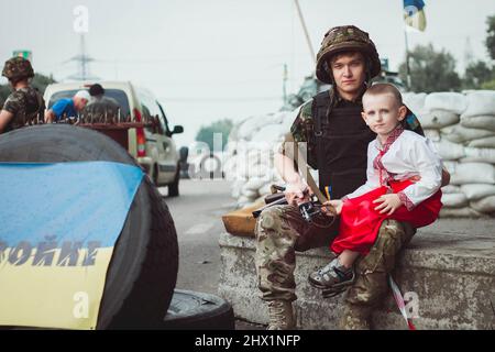 Ukrainischer Junge in traditioneller Nationaltracht sitzt in der Nähe eines ukrainischen Soldaten auf einer Straßensperre vor dem Hintergrund von Sandsäcken. Konzept des russischen Militärs Stockfoto