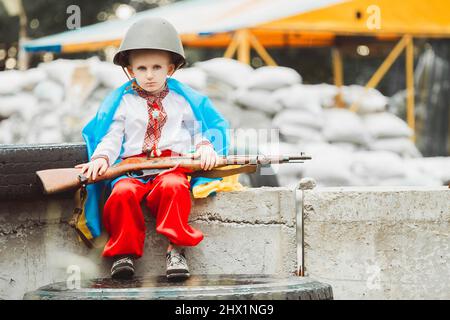 Ukrainischer Junge in traditioneller Nationaltracht und Helm sitzt auf einer Straßensperre, umwickelt in eine Flagge, mit Gewehr in den Händen vor Sandsäcken. Stockfoto